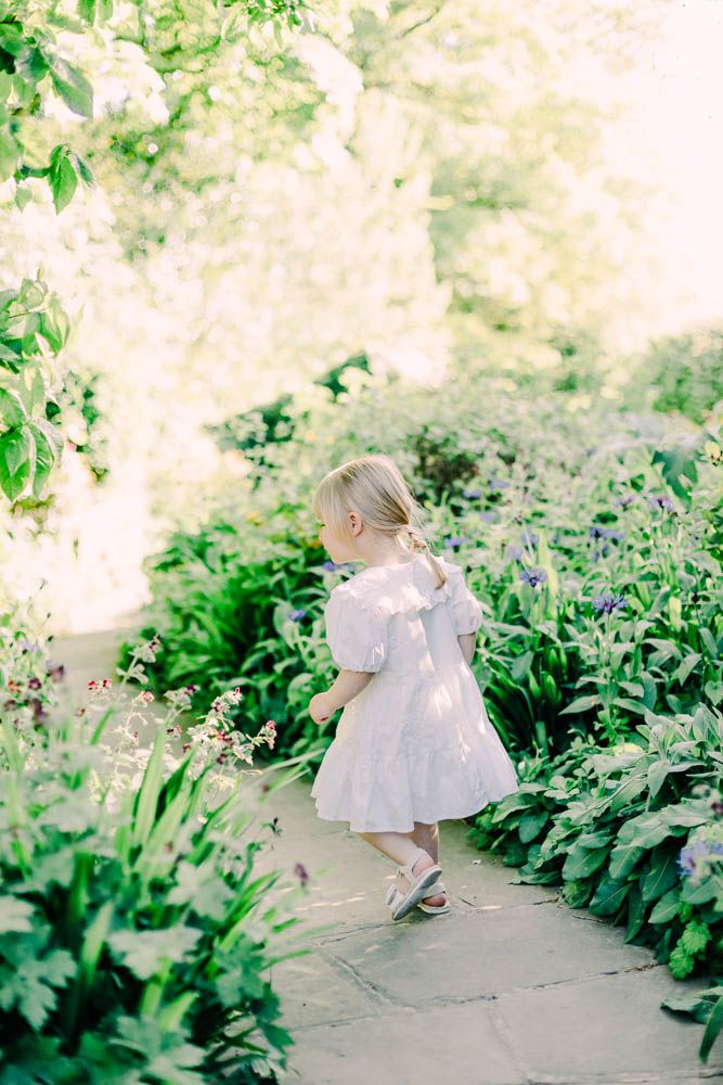 A small child in a white dress walks along a garden path surrounded by lush greenery and blooming flowers.