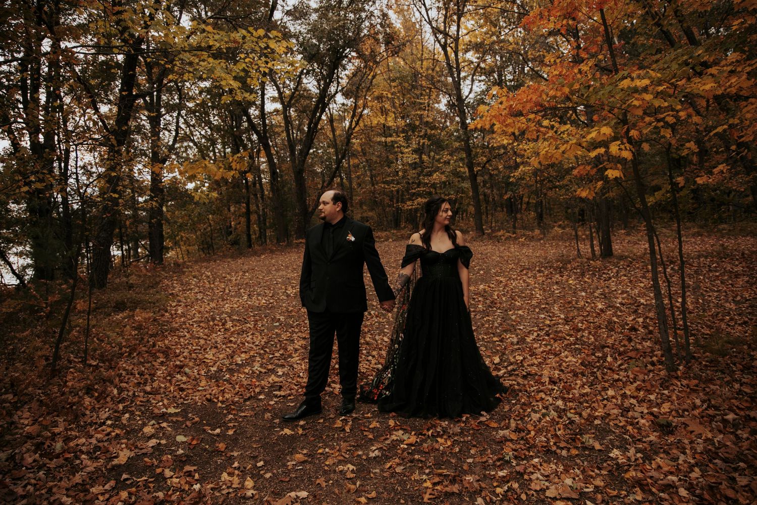 A couple in dark attire standing in a forest filled with fallen autumn leaves on the ground.