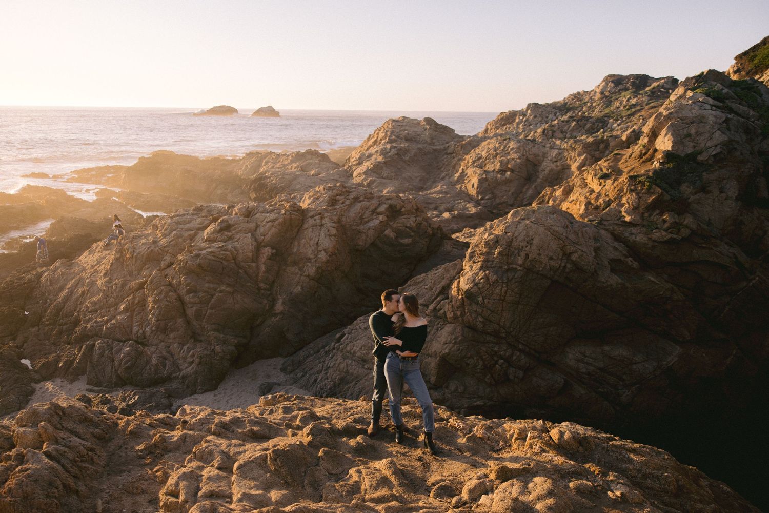 A couple silhouetted against golden sunset light embraces on rocky coastal cliffs overlooking the ocean.