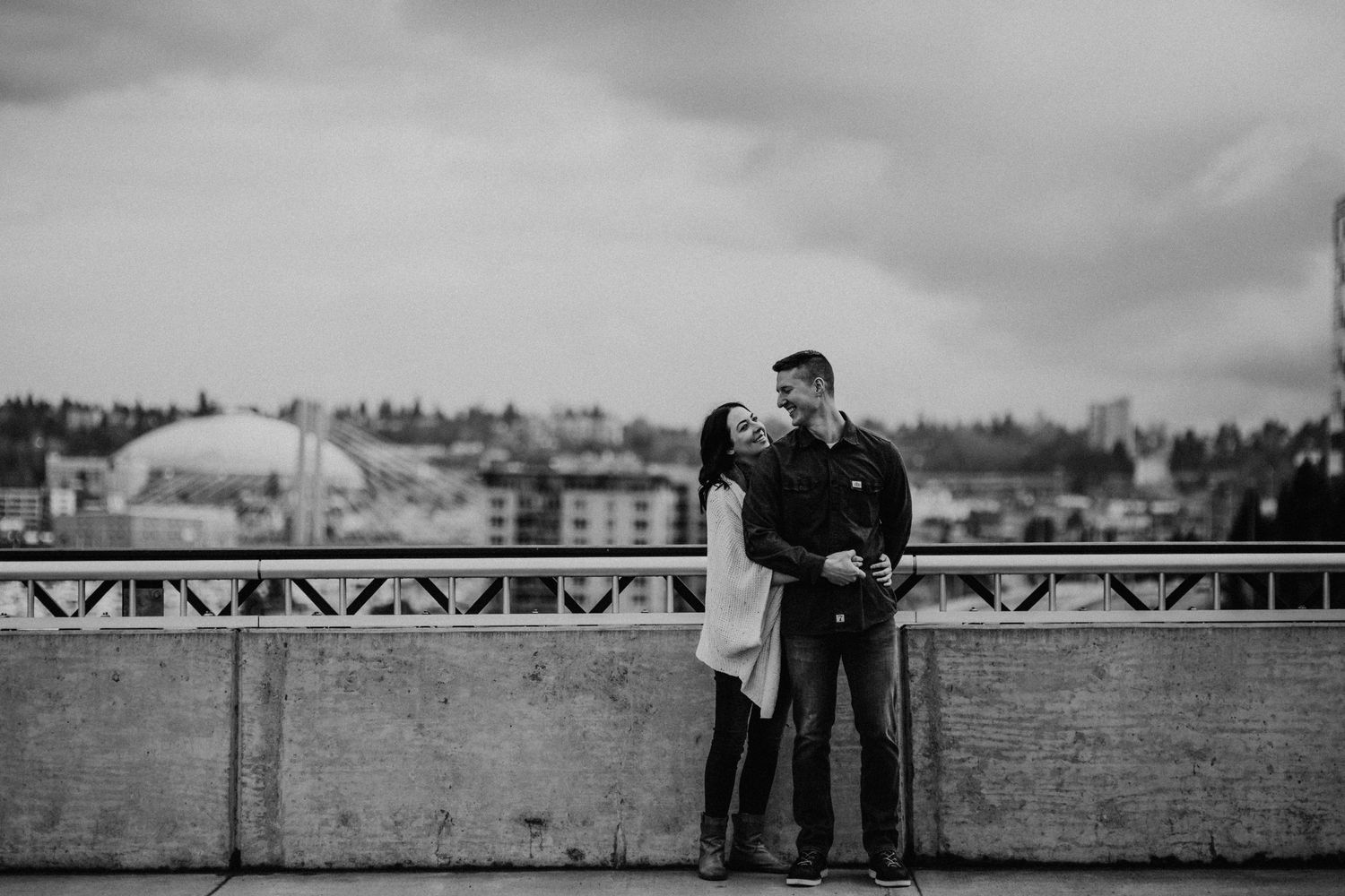 A couple stands close together on a bridge during an overcast day in black and white photography.