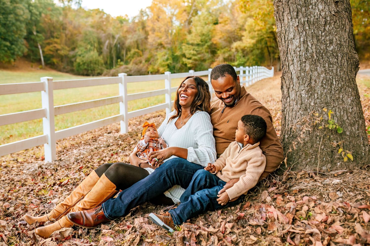 Family enjoying autumn day together sitting on fallen leaves by white fence.