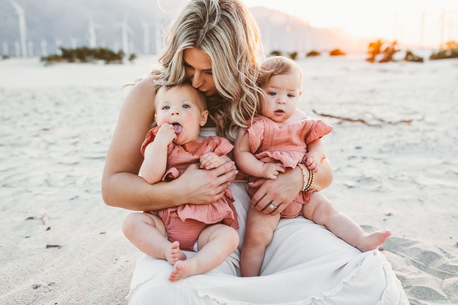 A mother of twins girls sits in the sand in palm springs, ca kissing her daughter's head