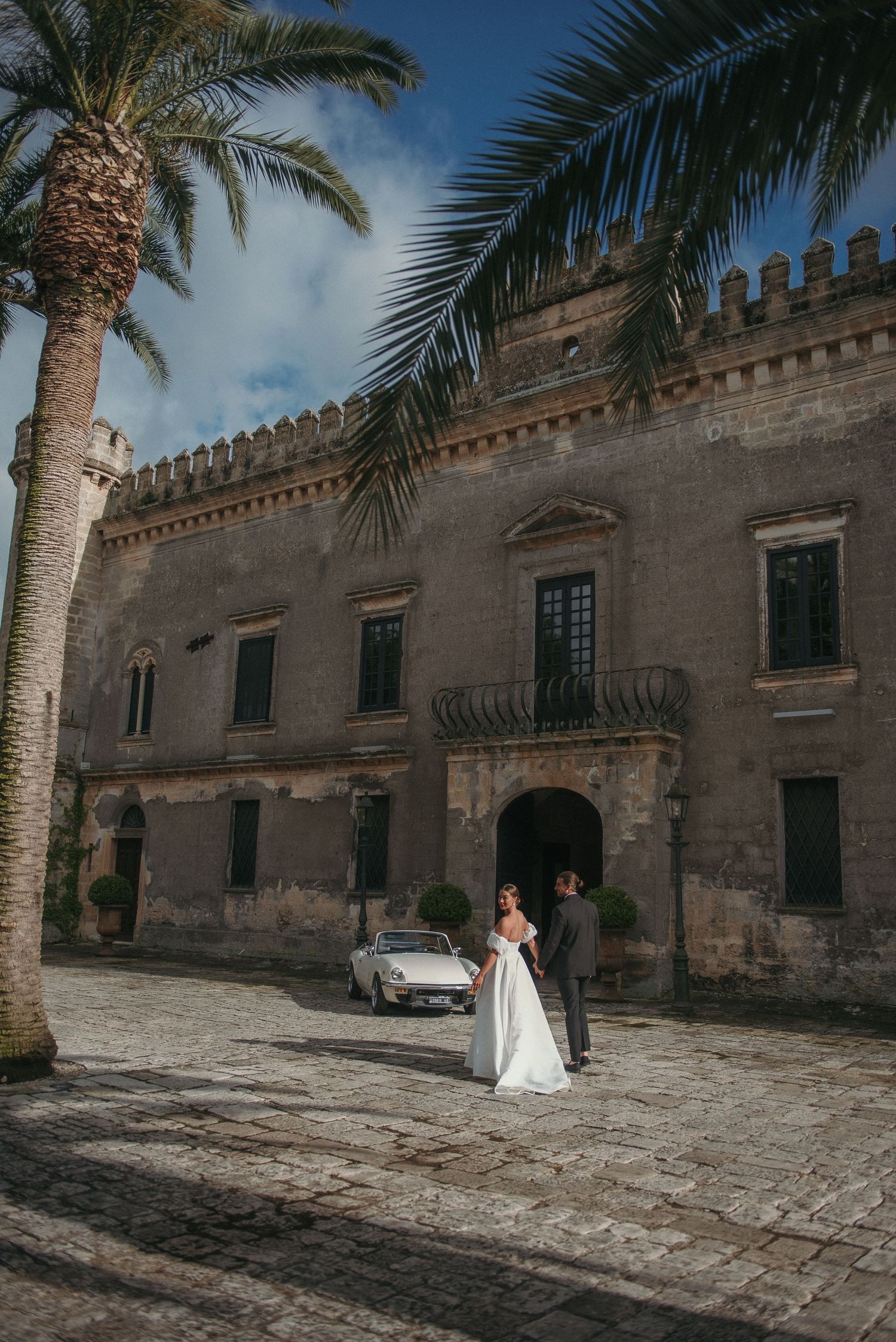 Wedding celebration at historic stone castle with palm trees and vintage car in Sicily, Italy.