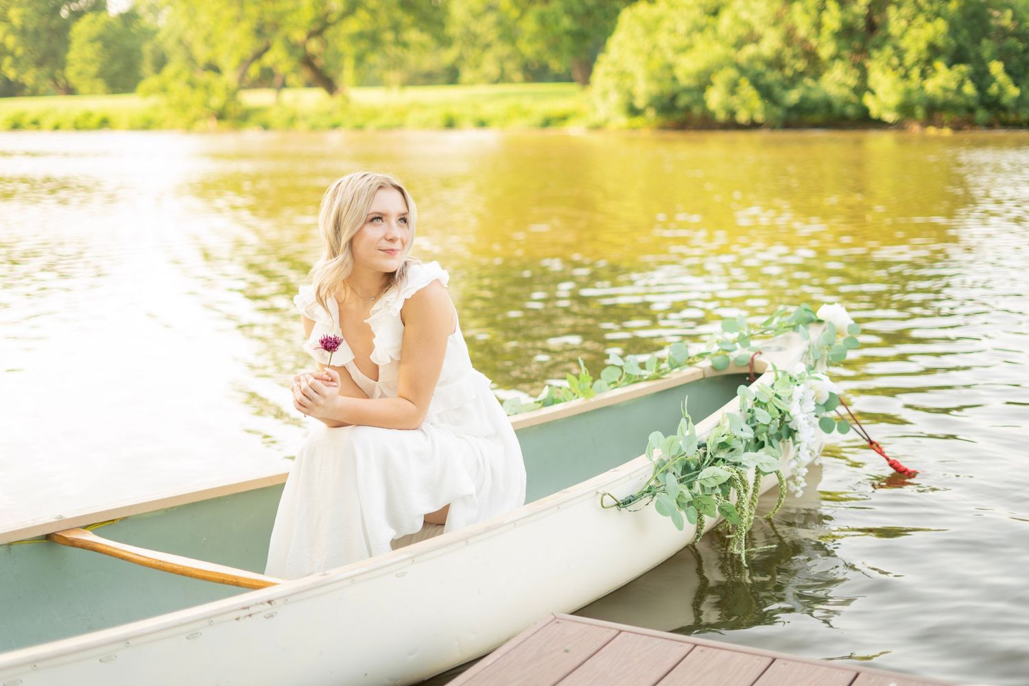 Blonde woman in white dress sitting in a wooden canoe on a sunlit lake holding a rose.