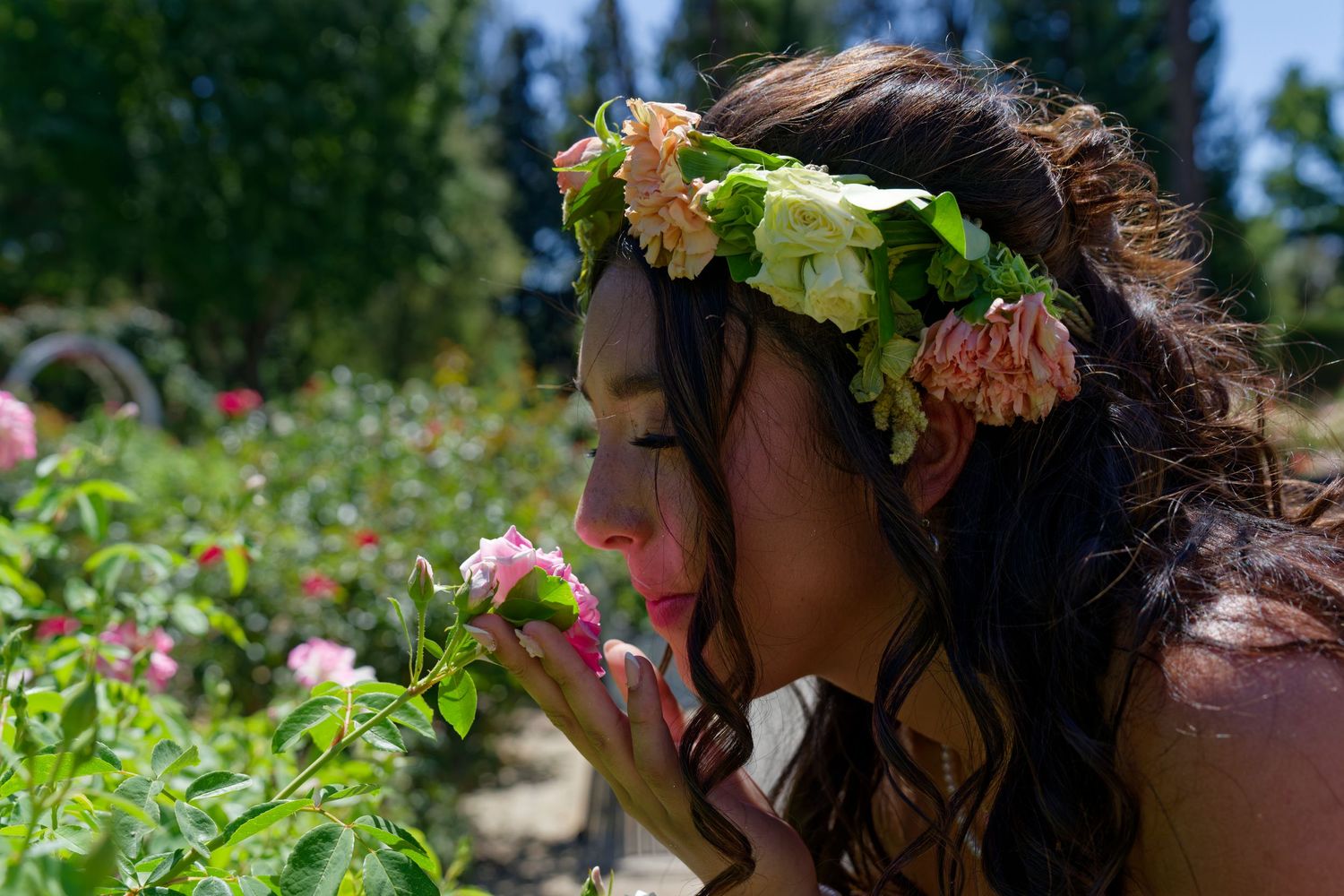 Young woman with floral crown smelling a pink rose in a sunny rose garden.