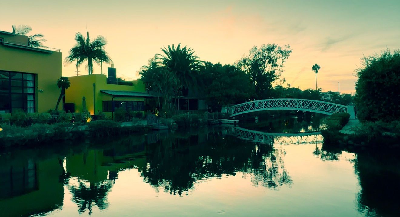 Serene Venice Canal at dusk with a decorative bridge, lush palm trees, and colorful reflections on calm water.