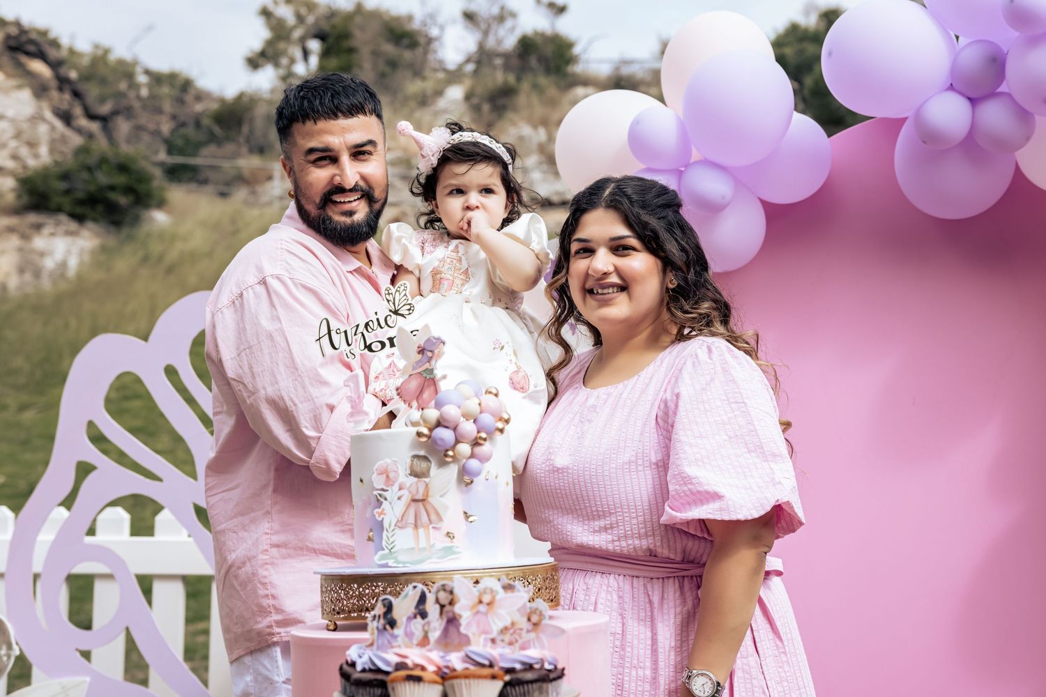 Family celebrating baby girl's first birthday with pink cake and balloon decorations outdoors.