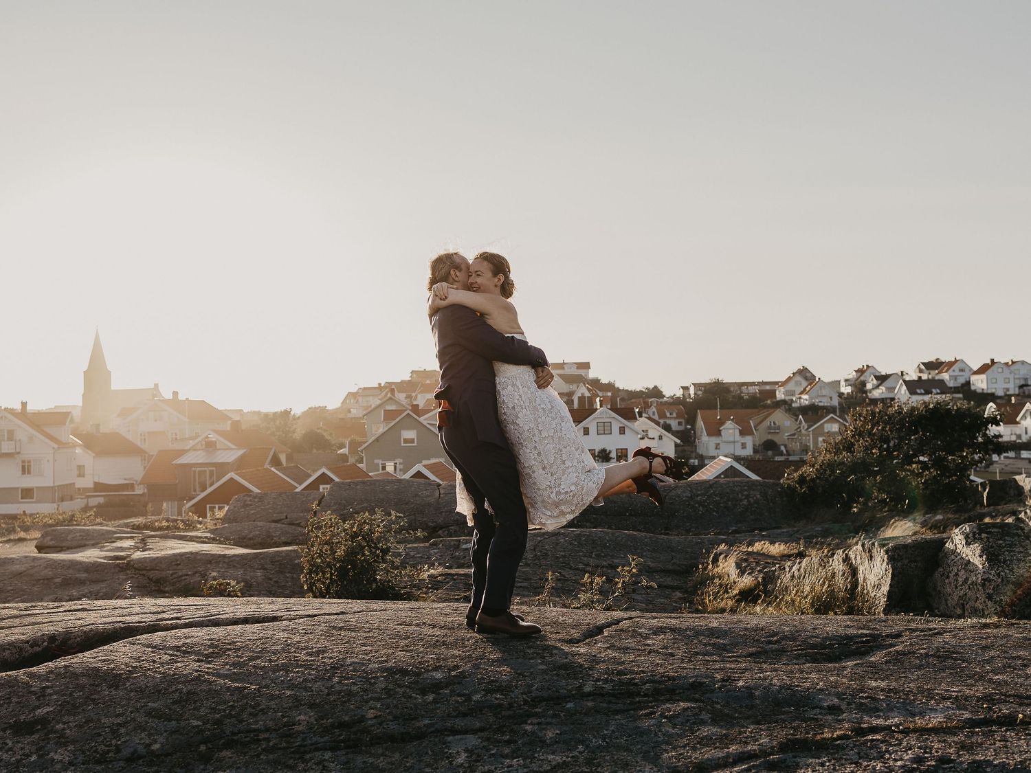 Groom lifting bride on rocky coastal landscape with Scandinavian village and golden sunset light in background.