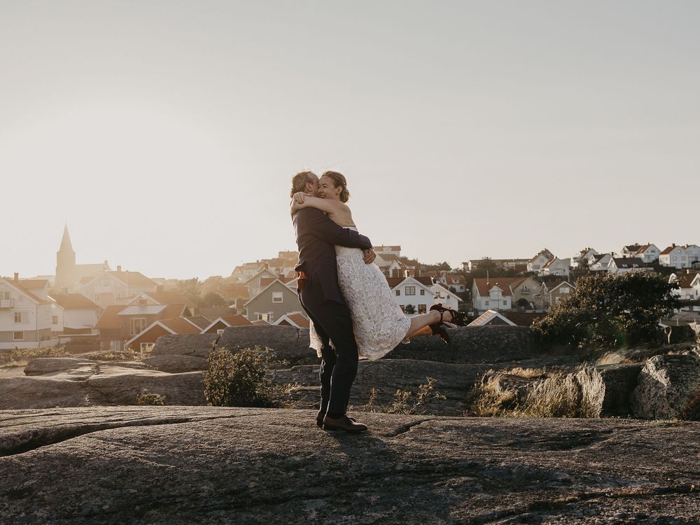 Groom lifting bride on rocky coastal landscape with Scandinavian village and golden sunset light in background.