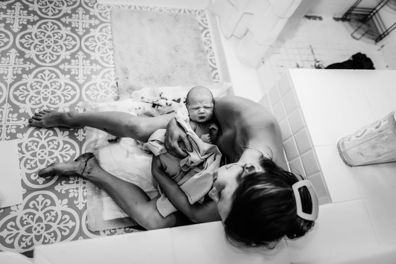 Black and white overhead photo of a mother resting on a patterned tile floor, cradling her newborn baby after a home birth.