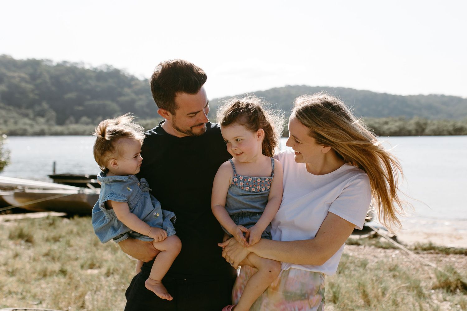 Happy family of four smiling outdoors near a lake, parents holding two young daughters on a sunny day.