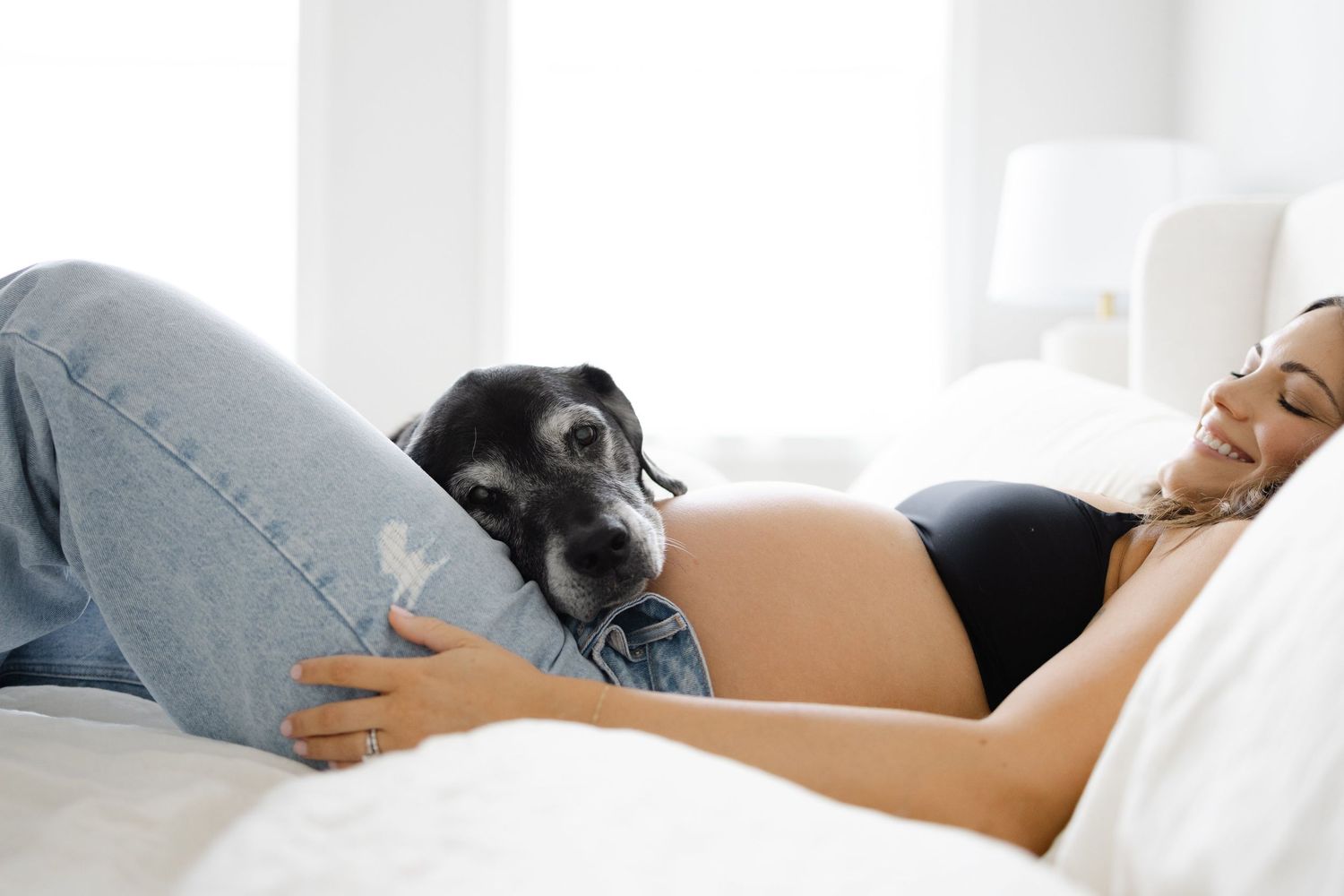 Pregnant woman lying on white bed in black crop top and jeans, cuddling with a black dog on her baby bump.