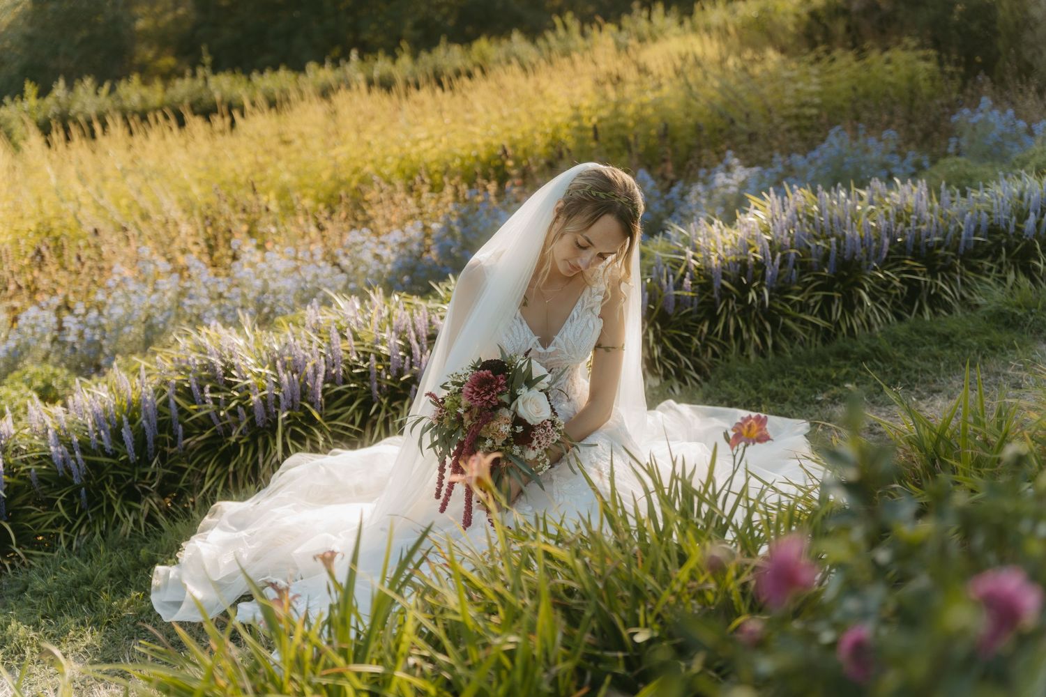Bride in white gown and veil sits in a lush flower field holding a bouquet, bathed in warm golden sunlight.