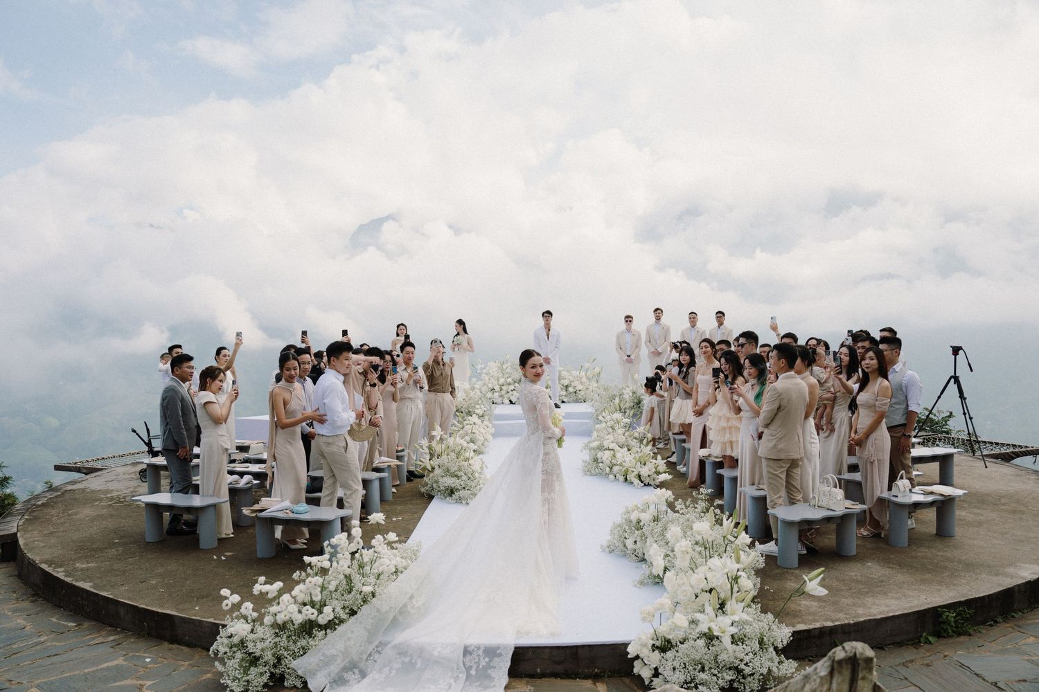 Wedding guests gathered on a beach dock during sunset celebration with white floral decorations and ocean views.