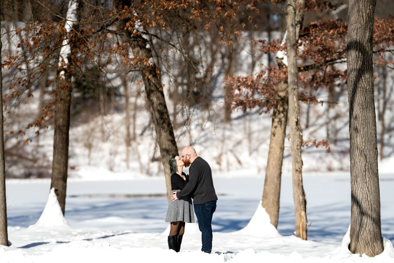 A couple shares a romantic kiss surrounded by snow-covered ground and bare trees in a winter forest setting.