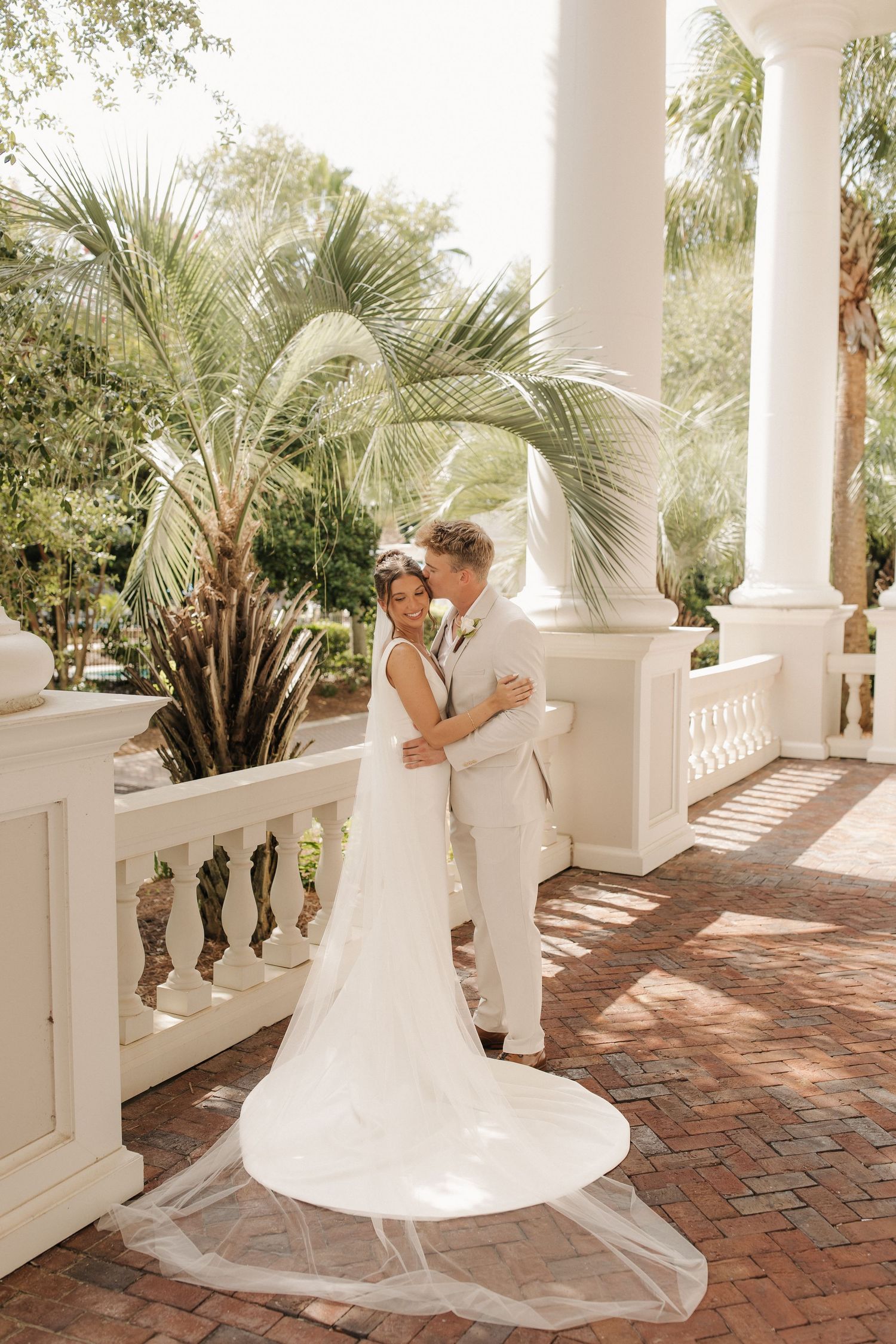Romantic wedding couple shares tender moments on a brick patio with white columns and tropical palm trees.