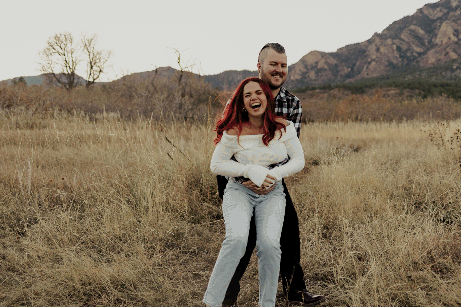 A couple embraces and laughs together in a golden field with mountains in the background during sunset.