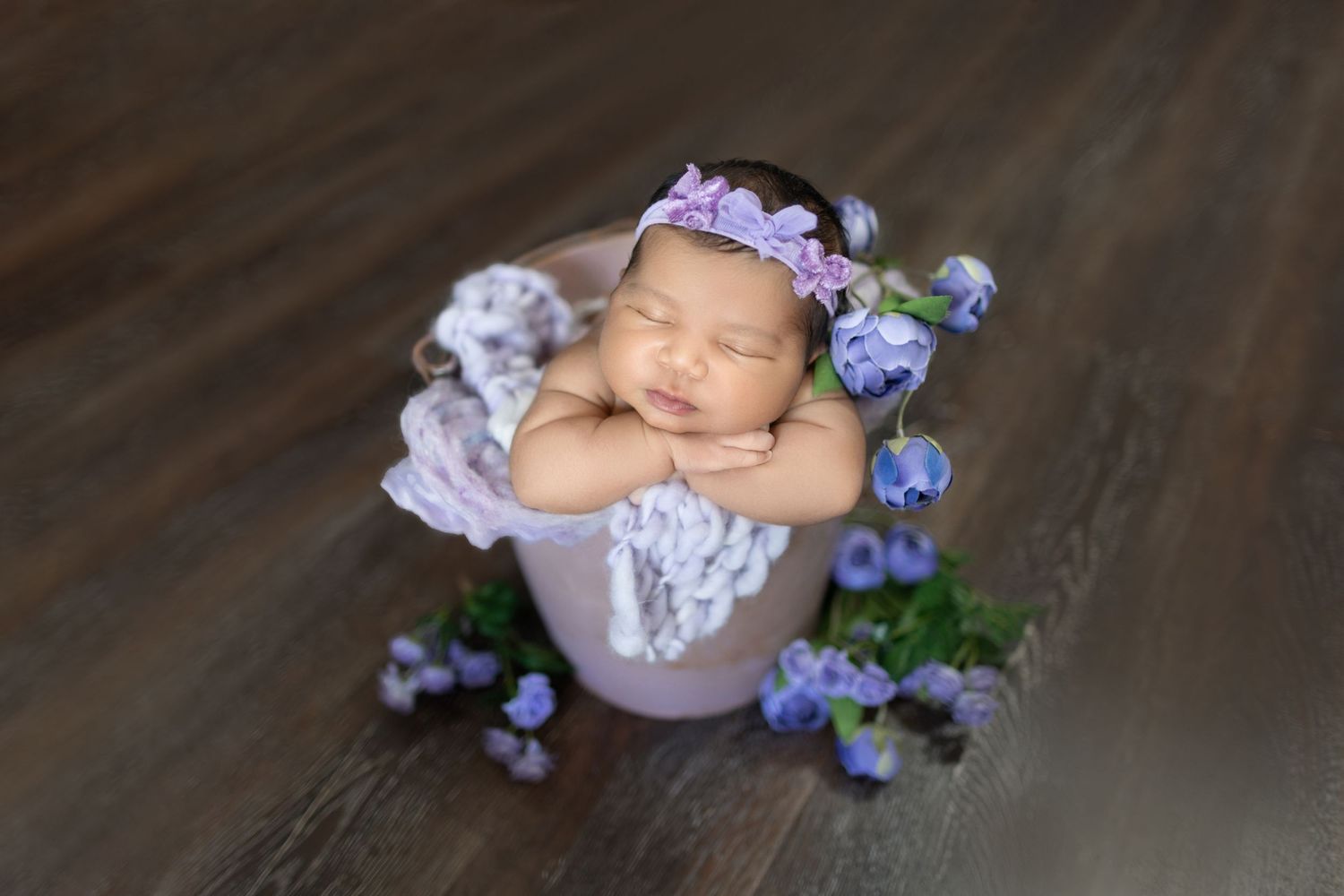 Sleeping newborn baby posed with purple flowers and soft white wraps on wooden backdrop.