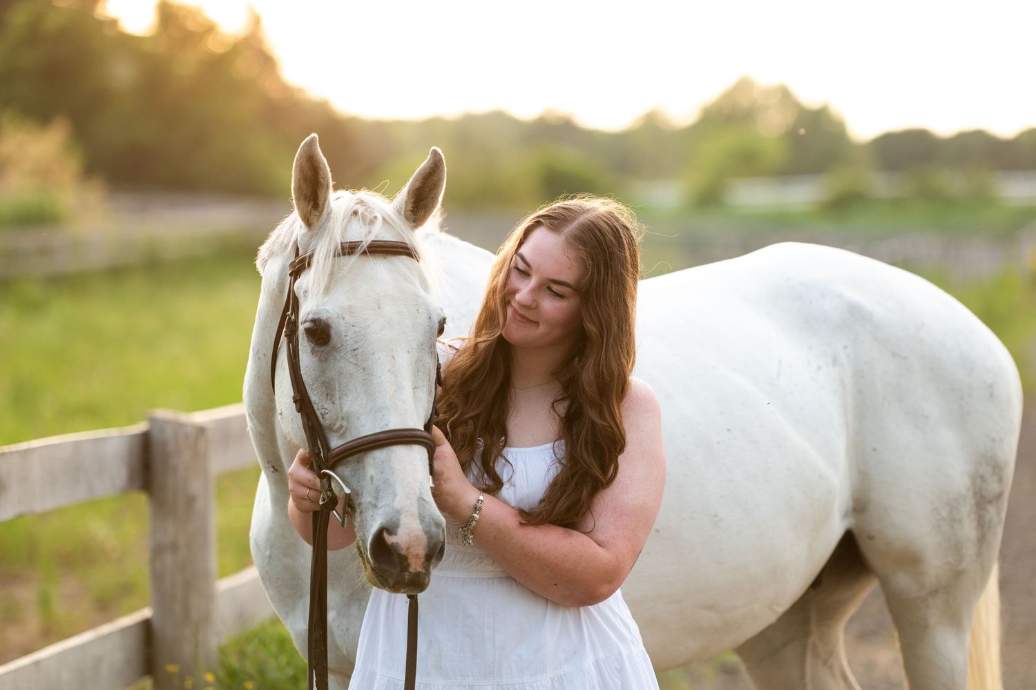 Young woman in flowing dress poses with beautiful white horse in sunlit meadow during golden hour.