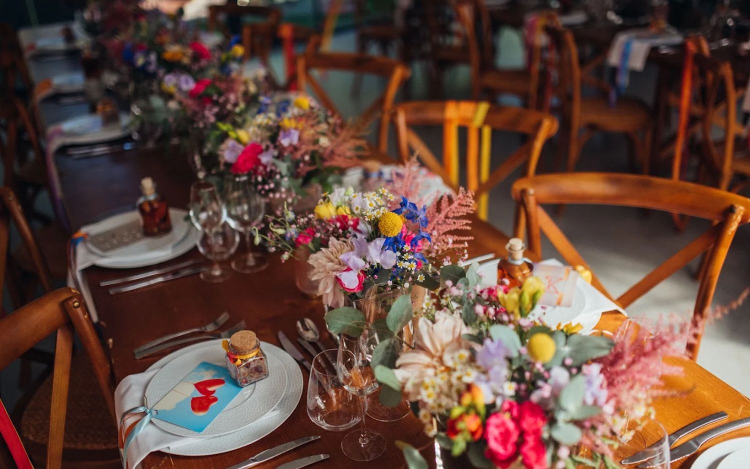 Rustic wedding reception table settings with colorful floral centerpieces and wooden chairs in warm lighting.