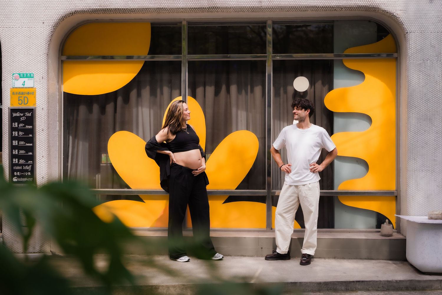 People sit on a concrete bench against a yellow wall with a curved design pattern in an urban setting.