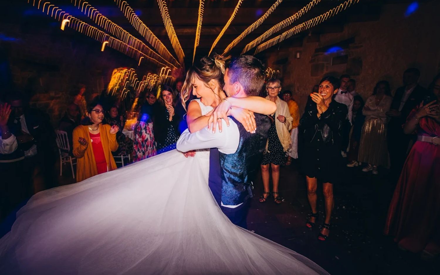 A group of people dancing and celebrating at a lively wedding reception with colorful lighting and a flowing white dress.