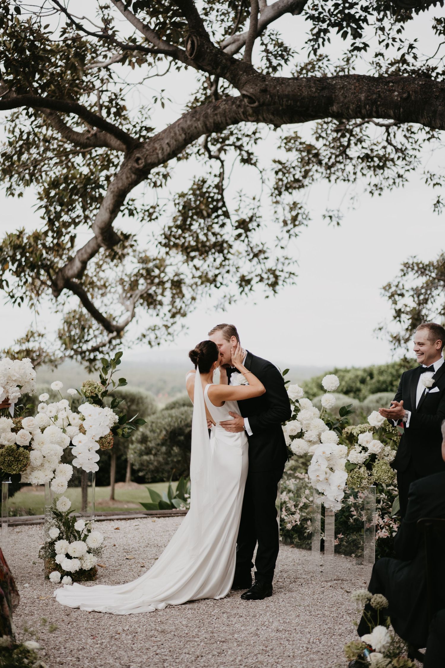 A romantic wedding couple embraces on a tree-lined gravel path decorated with elegant white floral arrangements.