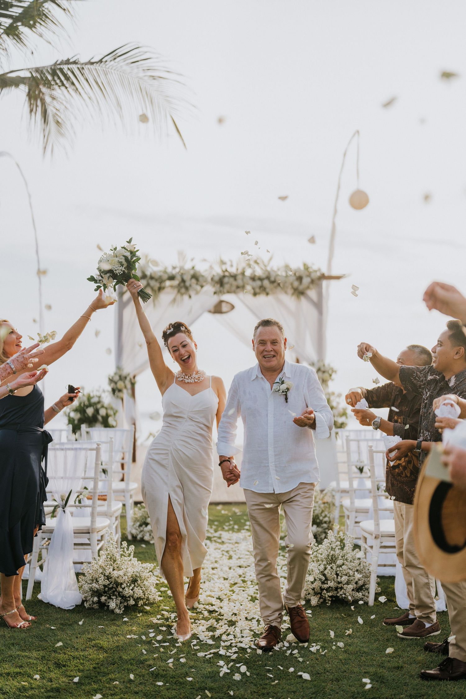 Wedding guests celebrate joyfully by tossing flower petals in the air at an outdoor ceremony.