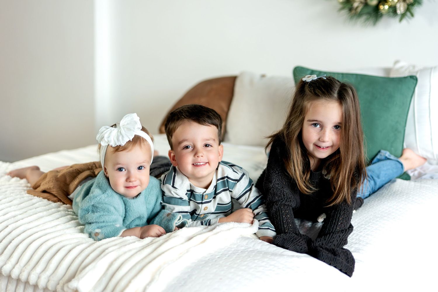 Three young siblings lying on a white bed with holiday decor in a cozy Christmas setting.