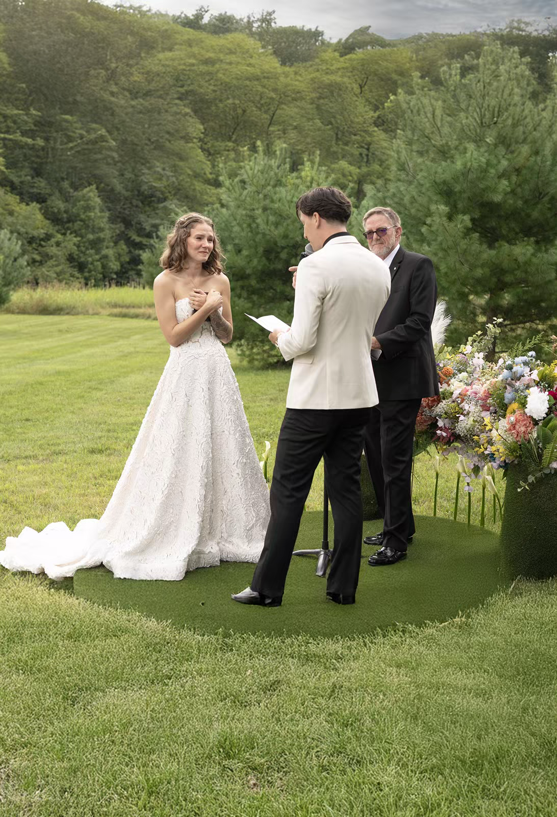 A wedding ceremony takes place outdoors on a lush green lawn with floral arrangements and trees in the background.