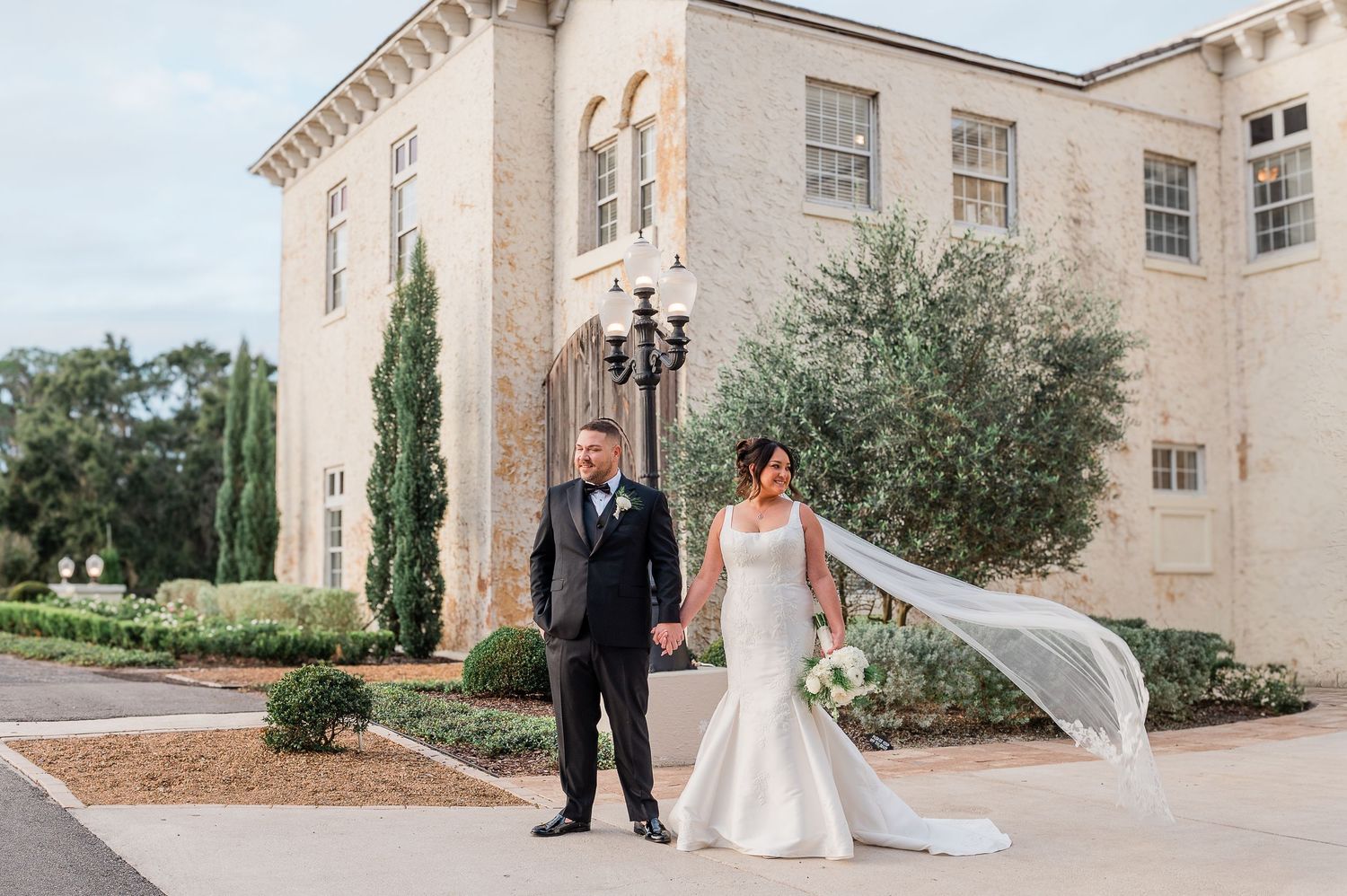 A wedding couple stands in front of a grand Mediterranean-style villa with stone columns and arches.