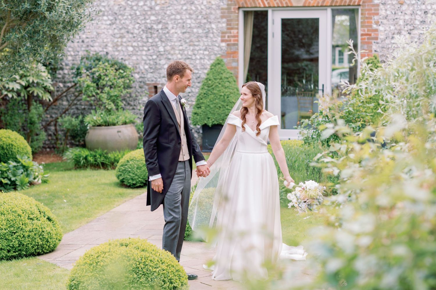 A couple strolls through a lush garden pathway beside a rustic brick wall and flowering plants on their wedding day.