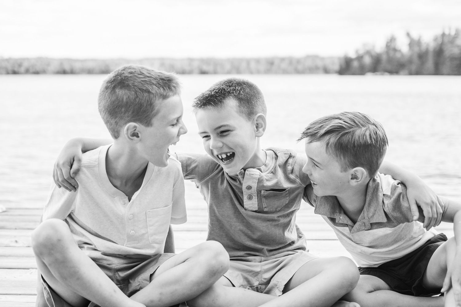 Three children share laughs while sitting together on a dock by the lake.