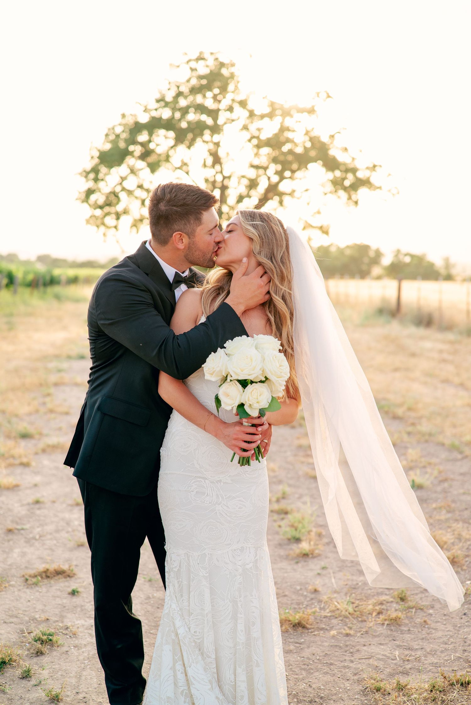 A romantic wedding couple shares a kiss while holding a white bouquet in an outdoor setting.
