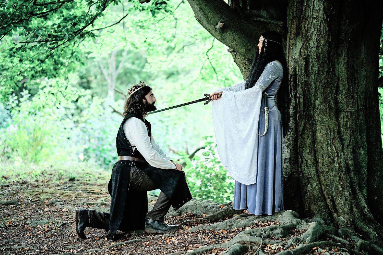 Two figures in white and black clothing sit beneath a large tree trunk in a lush green forest setting. LOTR maternity shoot.