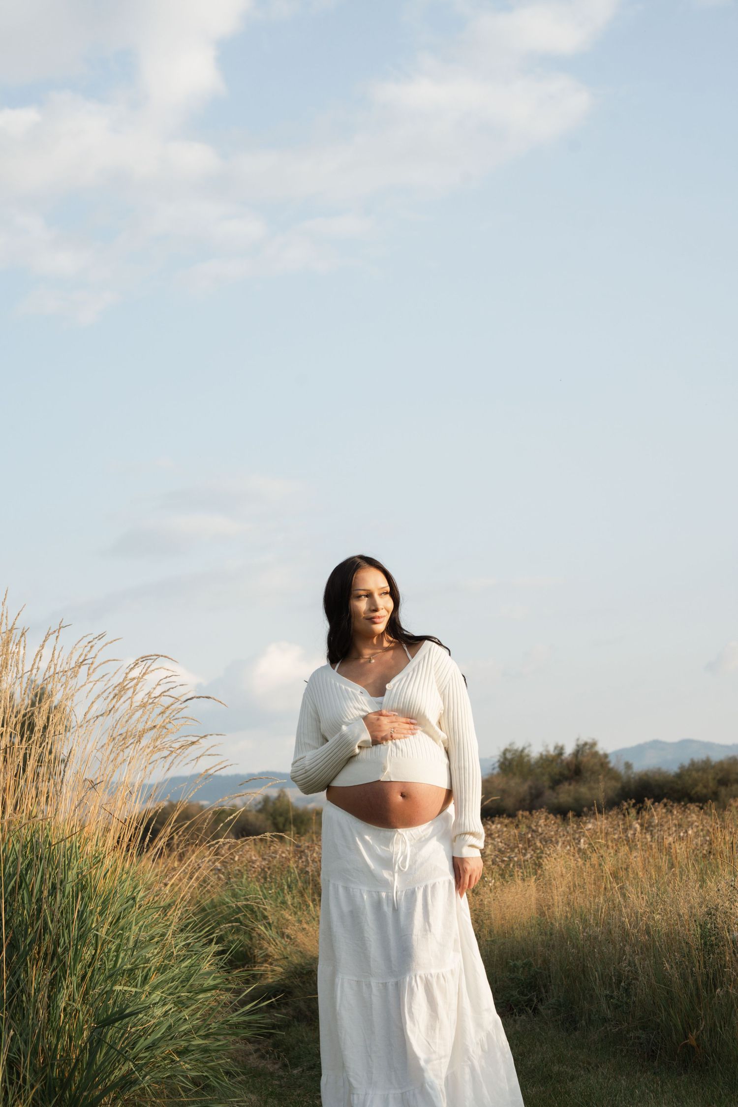 A pregnant woman in a white dress stands in a golden field at sunset with tall grass swaying in the breeze.