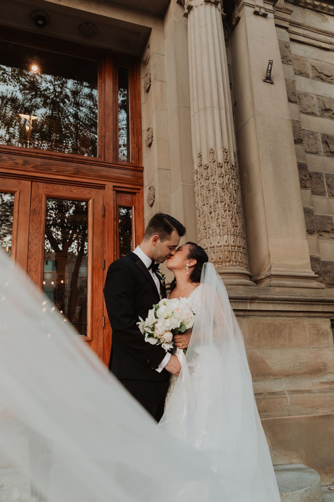 A bride and groom share a romantic kiss outside a grand stone church, her veil flowing in the wind.