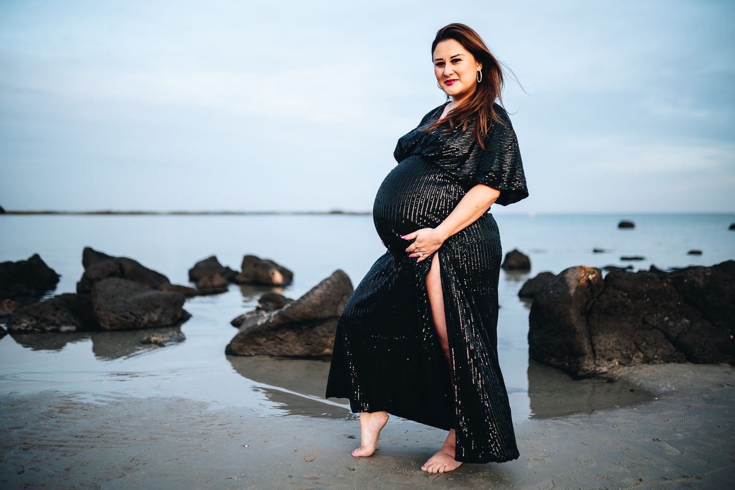 Pregnant woman in elegant black dress poses barefoot on rocky beach at twilight taken by Melbourne maternity photographer during in Wyndham