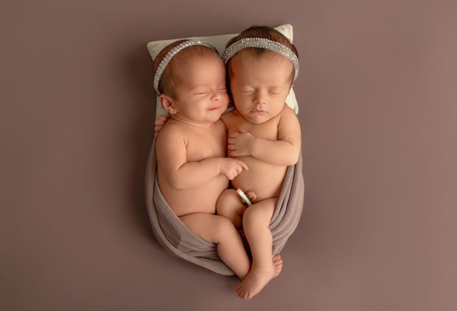 Two sleeping newborns in pink and purple outfits lay in individual white bowls surrounded by delicate floral decor.