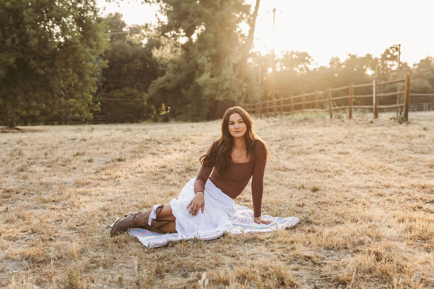 A person in a brown sweater sits on a blanket in a sunlit field during golden hour.