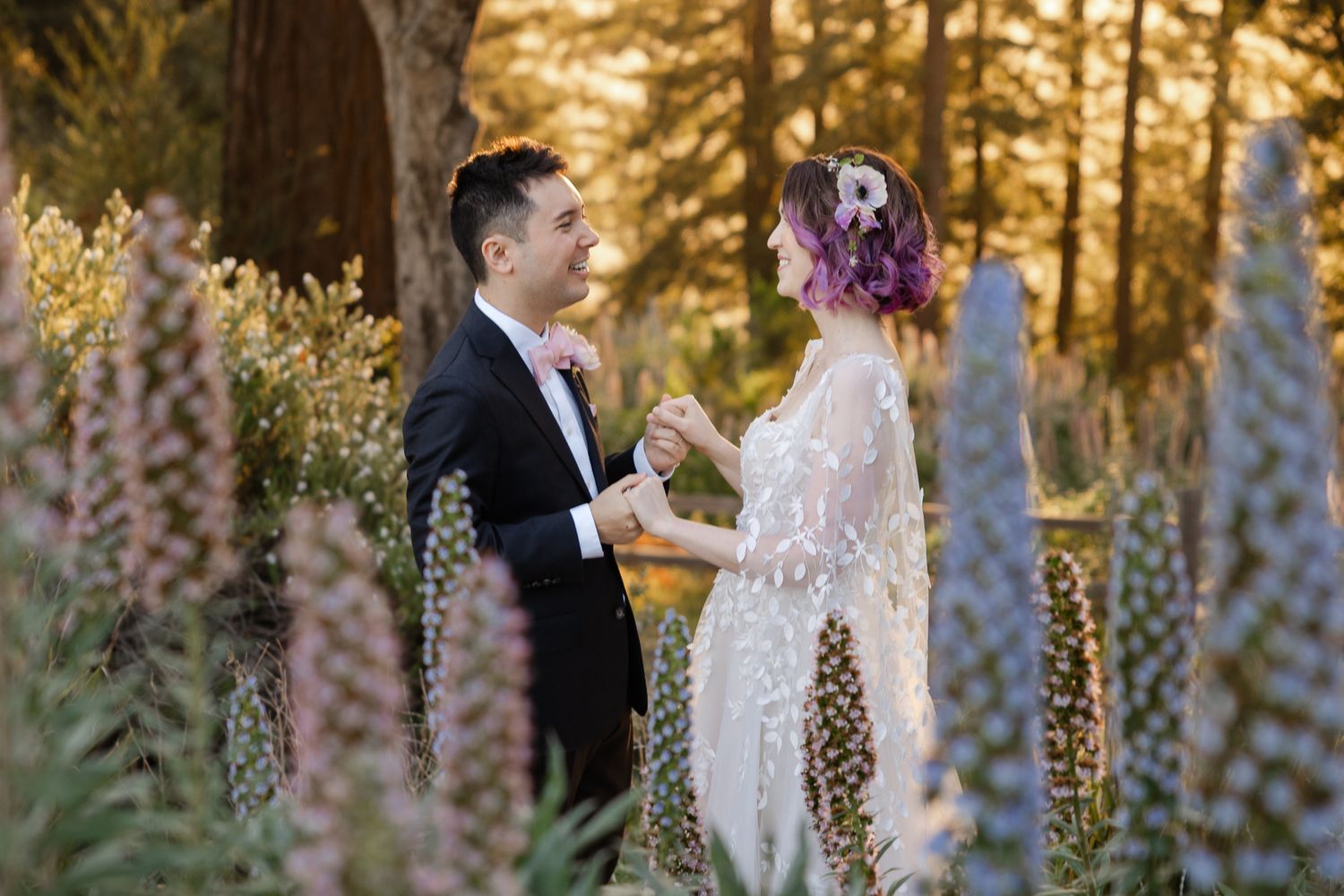 A couple stands among tall purple lupine flowers in a forest at golden hour during their wedding ceremony.