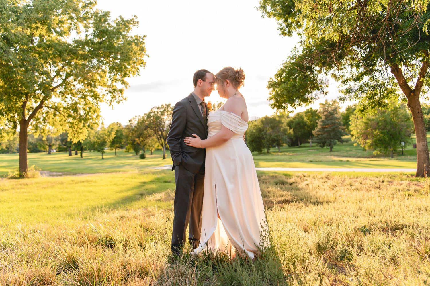 A couple embraces in a golden sunlit field during sunset, surrounded by trees and tall grass.