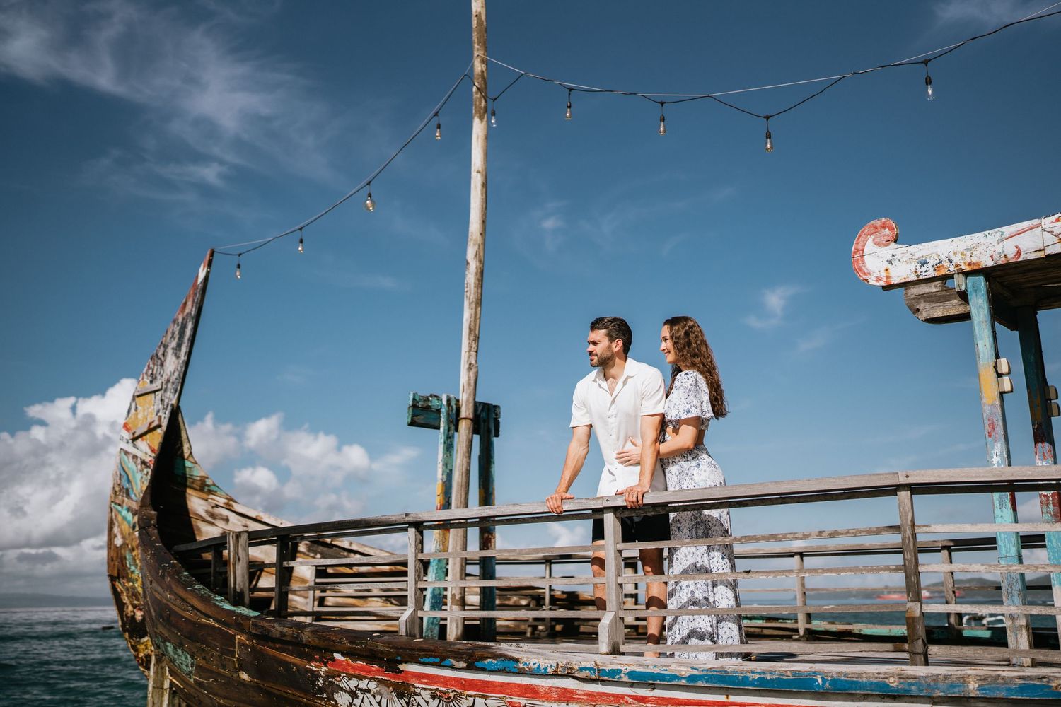 A couple stands together on a wooden pier with string lights and blue skies overhead.