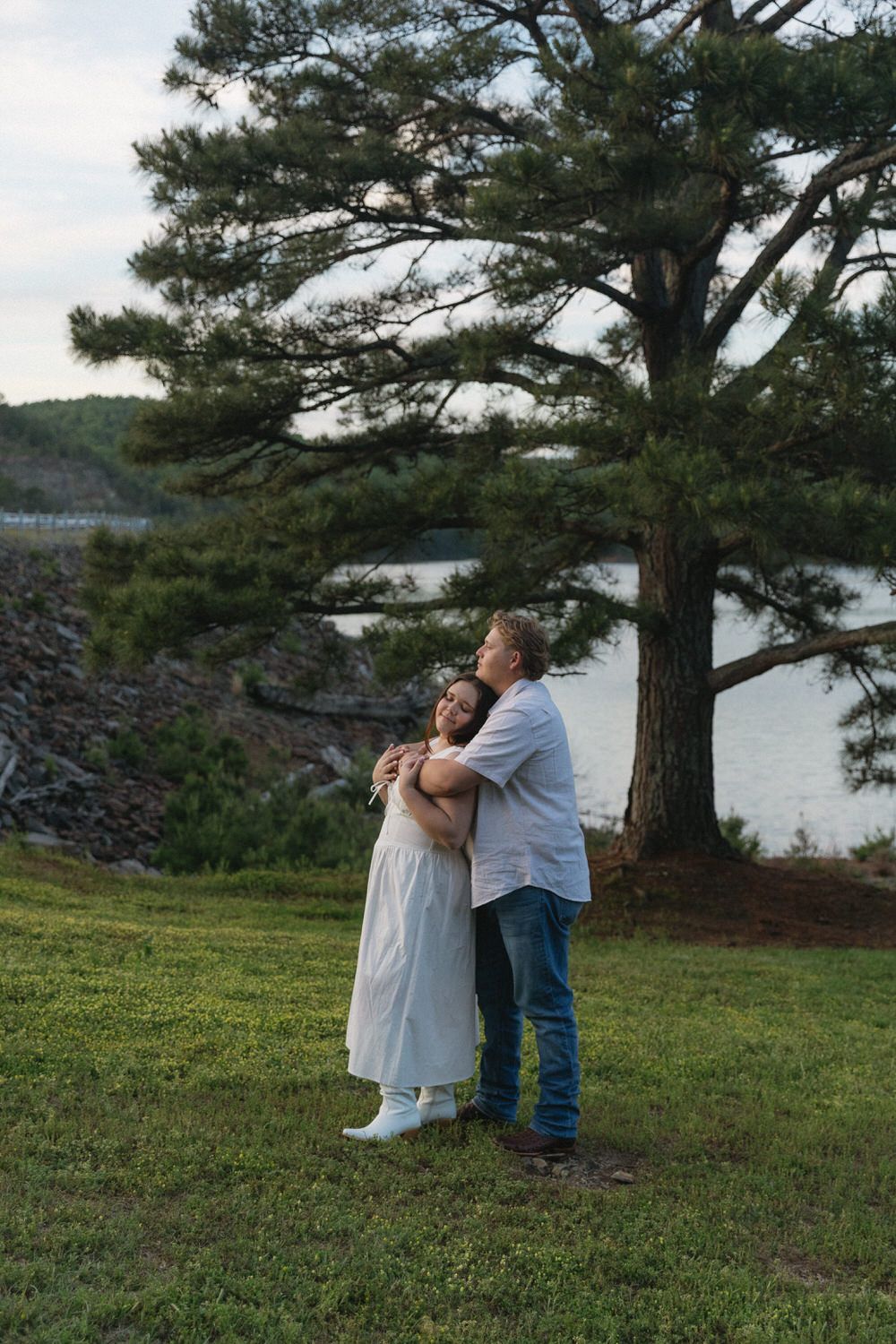 A couple embraces on a grassy hill overlooking Carter's lake with a tall pine tree in the background in North Georgia