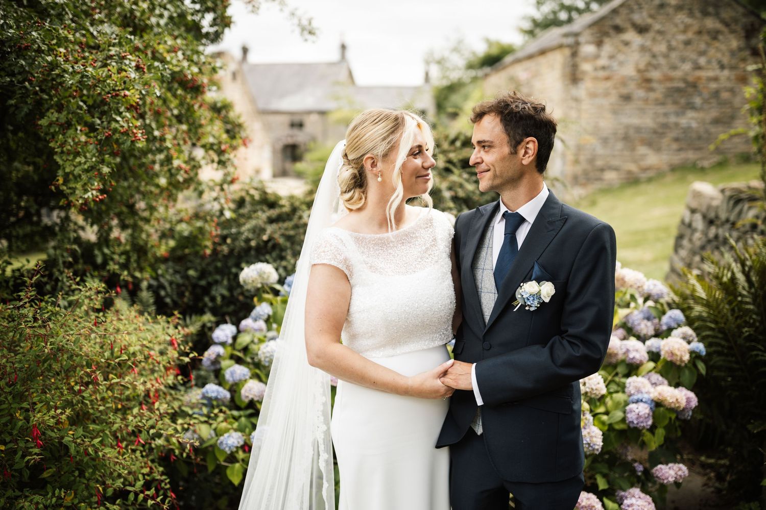 Couple shares romantic moments in garden wedding setting with stone walls and lush greenery in background.