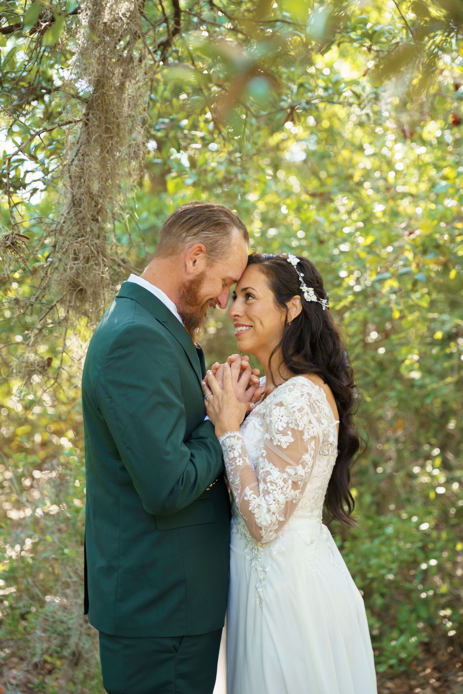 A romantic wedding couple embraces in a sun-dappled forest setting wearing green and white attire.