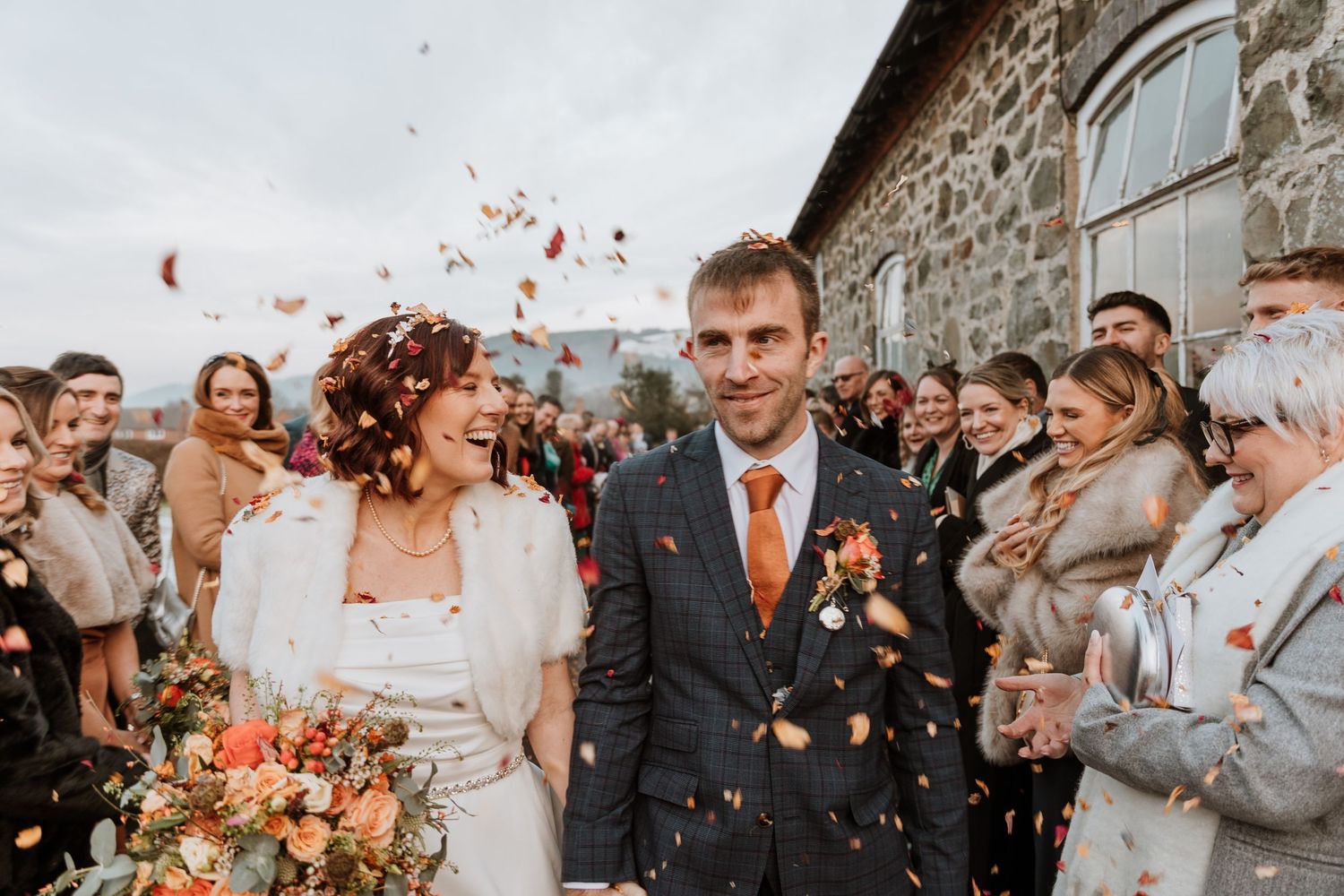 Newlyweds walk through shower of confetti outside stone building while wedding guests celebrate their special day.