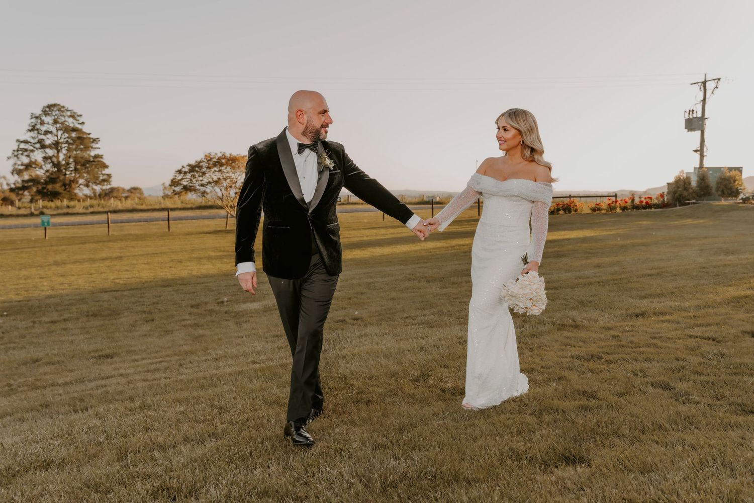 A bride in an off-shoulder white gown and groom in black suit hold hands on urban steps near vintage lamp post.
