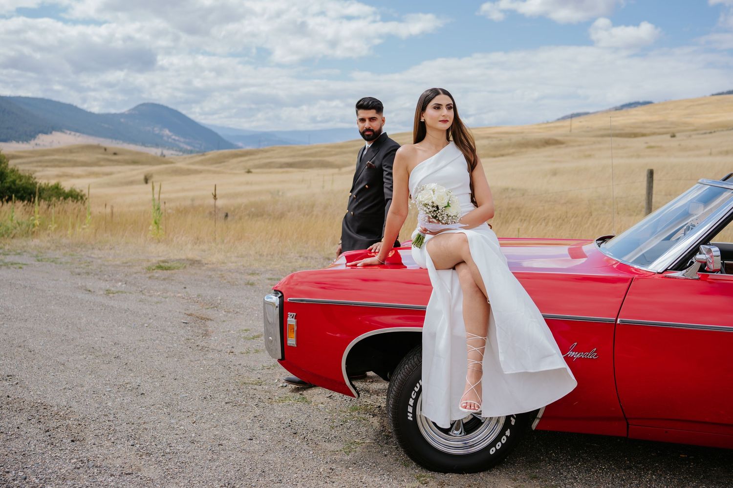 Couple poses with vintage red convertible car against scenic mountain landscape on their wedding day.