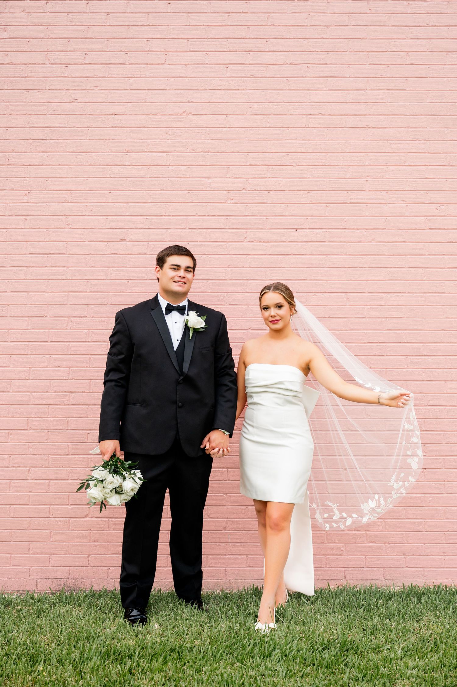 Couple in wedding attire poses playfully against pink brick wall during outdoor photo session.